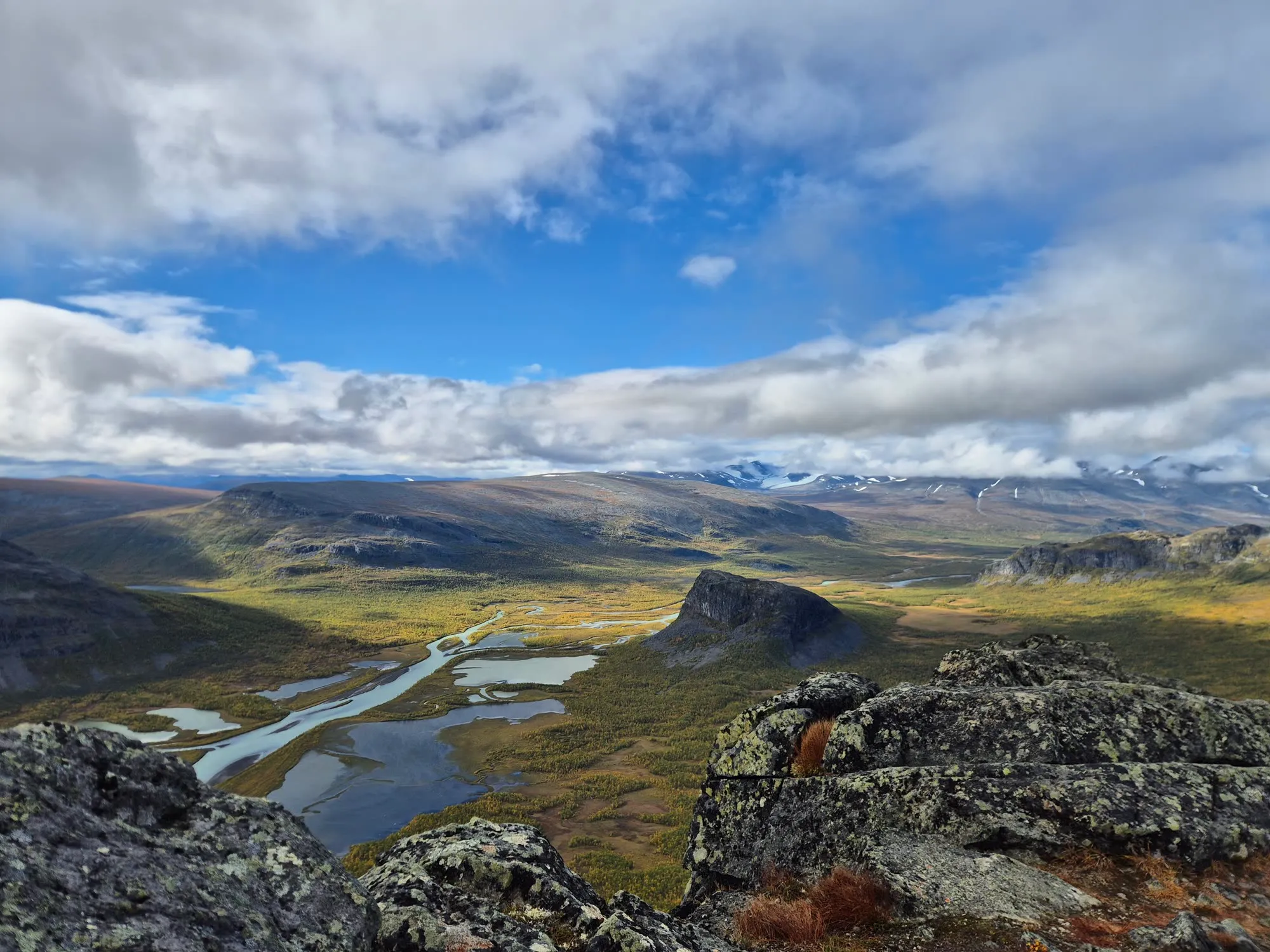 View from a rocky summit over a wide valley with a braided river, a lone flat-topped mountain and snowy peaks under clouds